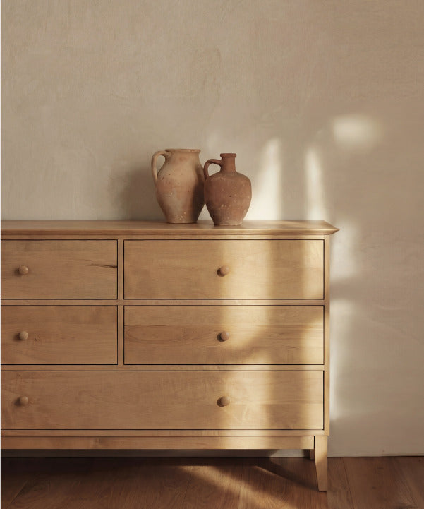 A dark brown wooden dresser with multiple drawers is centered against a white wall with decorative trim. Two mirrors and a vase with branches and books are displayed on the dresser.