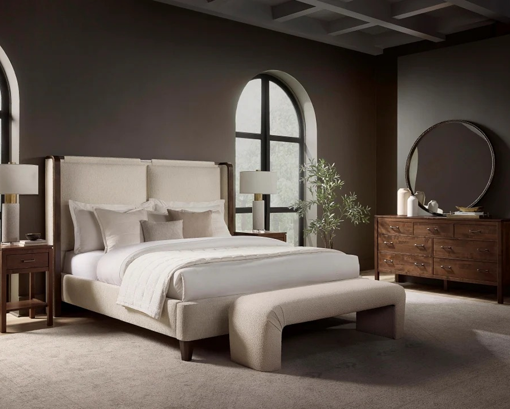 Elegant bedroom with a dark wooden bed, white and brown bedding, and matching side tables with lamps. A modern chandelier hangs above, and there’s a desk and chair to the left. The room has soft lighting and white walls.