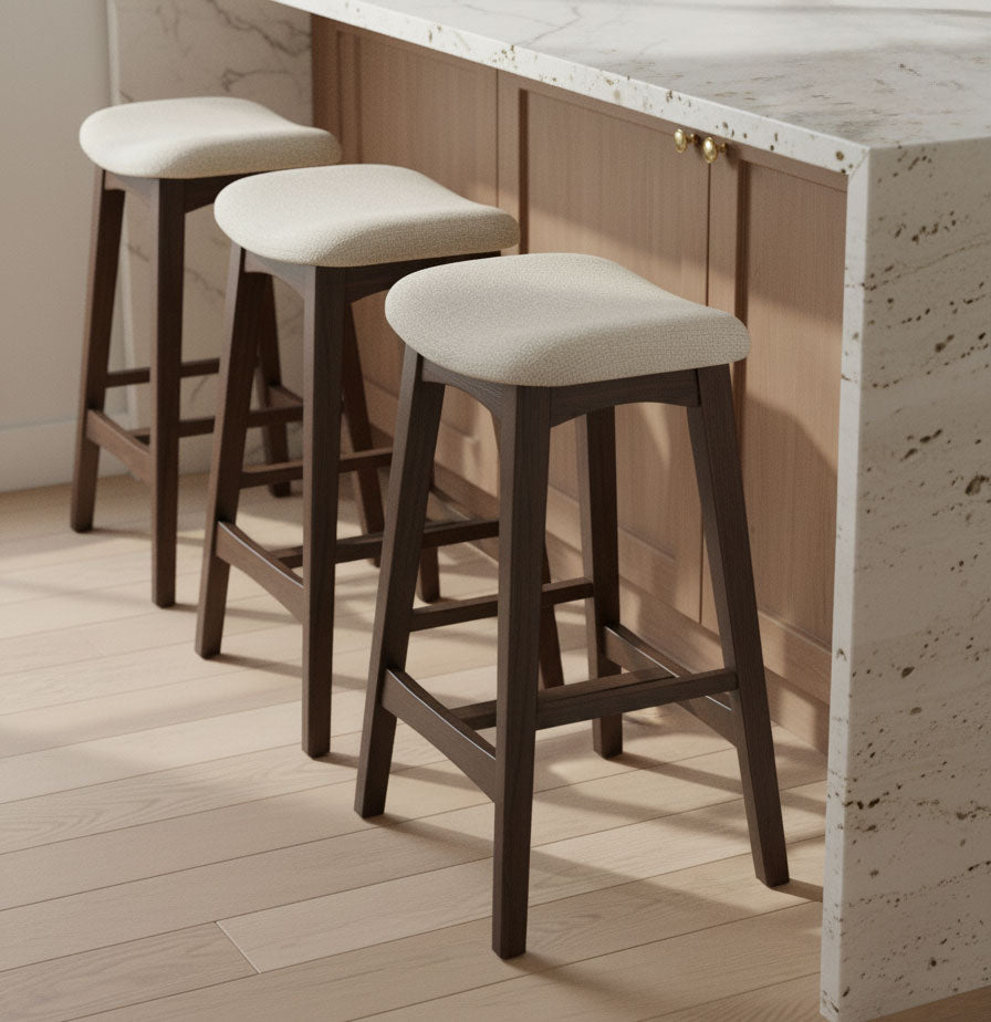Three wooden bar stools with light upholstered seats are lined up next to a marble kitchen island with light wood paneling. The stools are placed on a light-colored wooden floor.