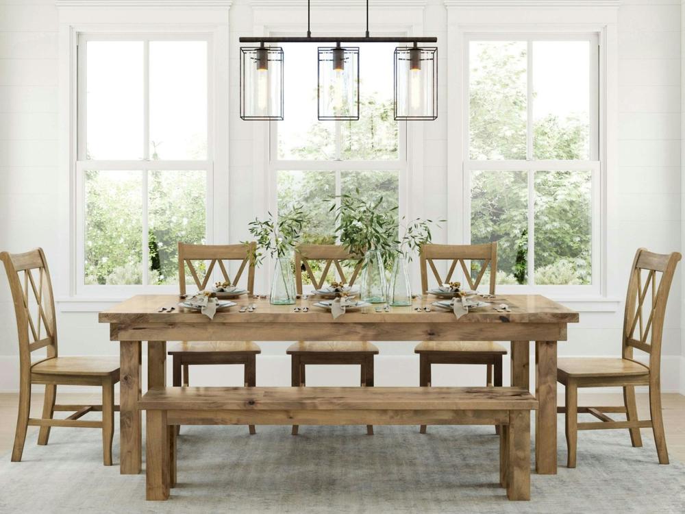 A James+James Farmhouse Bench in Harvest Wheat on Alder sits alongside a wooden dining table set for four with plates, cups, cutlery, and leafy green plants in glass vases by large windows. Four matching chairs complete the scene.