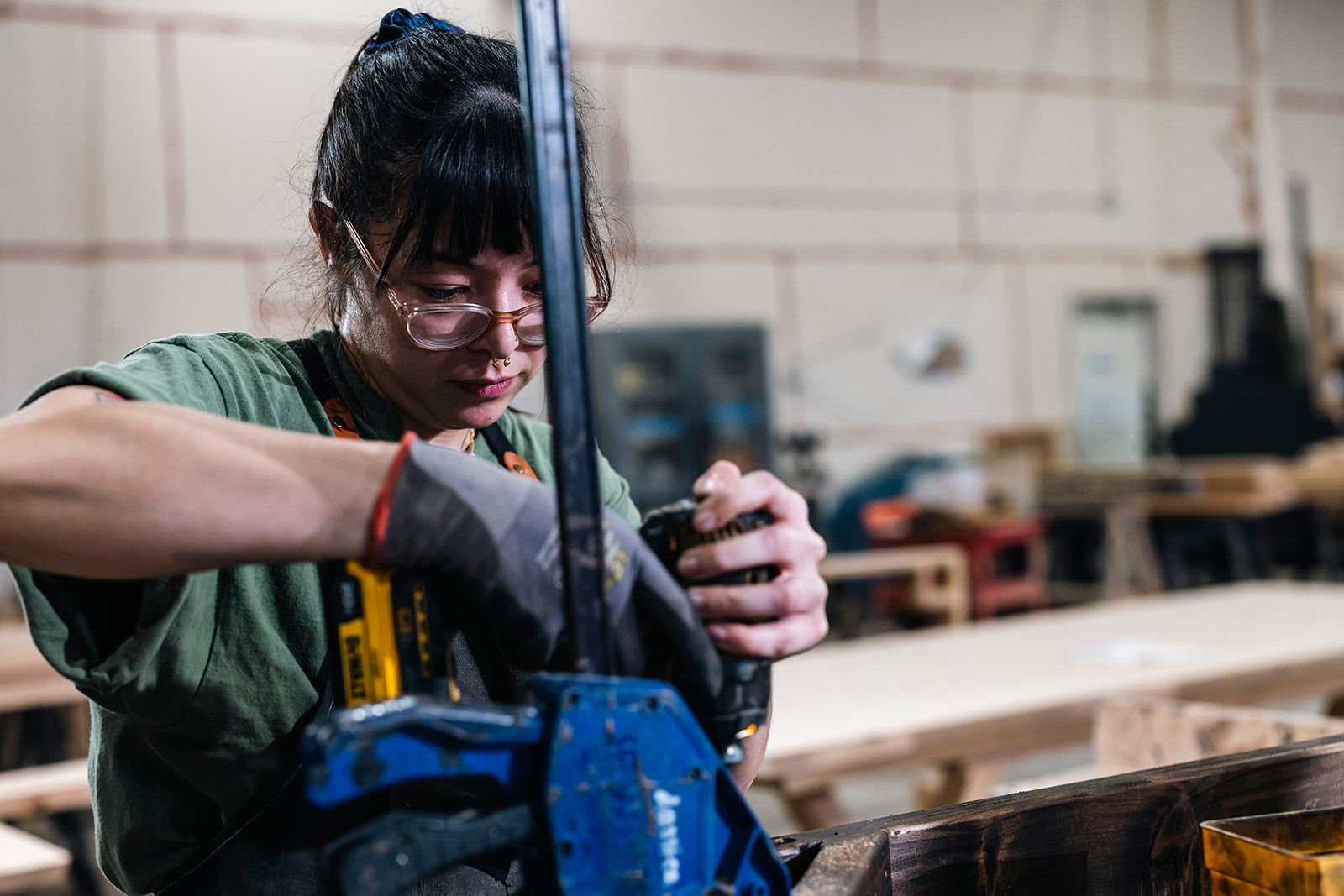 A person wearing safety glasses and gloves uses a large clamp on a wooden object in a workshop. They are focused on their task in an industrial setting with tools and materials in the background.