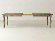 A French Country Expandable Dining Table in Harvest Wheat from James+James stands on a light-colored floor against a plain wall. The table features four turned legs and an extension mechanism in the middle with a visible wooden support system. The tabletop is split, showcasing the extended section.