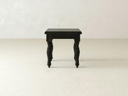 The Abigail End Table - Charred Ember by James+James, featuring ornate, turned legs in a black finish, stands on a plain, light-colored floor against a white wall. The table boasts a classic, vintage design with a smooth, rectangular surface.