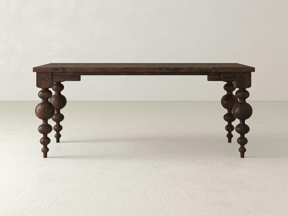 A dark wooden Olivia Desk from James+James, crafted from Knotty Alder, stands on a light, neutral-colored floor. The rectangular top is supported by four intricately carved Olivia Turned Legs, featuring a mix of spherical and cylindrical shapes stacked along their length.