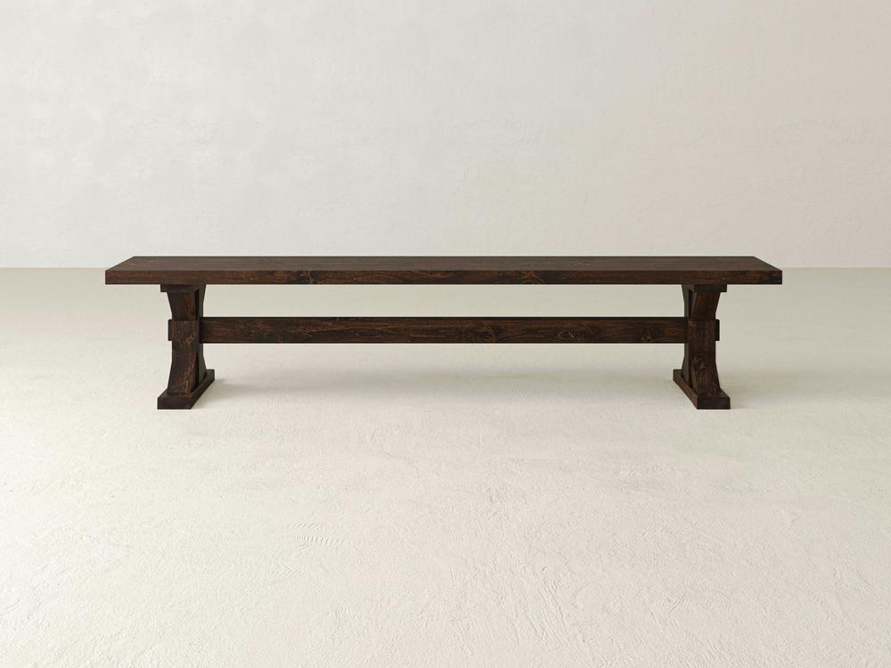 The James+James Trestle Bench in Tobacco on Alder features sturdy rectangular legs and a horizontal support beam, set against a light wall and floor.