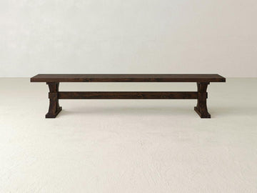 The James+James Trestle Bench in Tobacco on Alder features sturdy rectangular legs and a horizontal support beam, set against a light wall and floor.