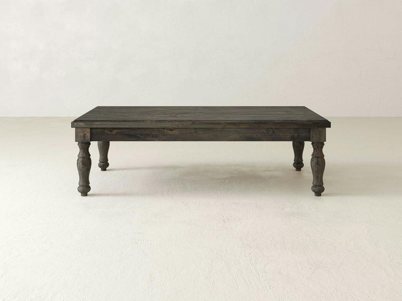 An Abigail Coffee Table by James+James, featuring a dark wooden rectangular design with four intricately carved legs, stands on a light-colored floor. The table boasts classic charm with its rustic, vintage appearance and handcrafted solid wood construction. The background is plain with a slightly textured off-white wall.