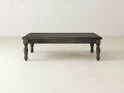 An Abigail Coffee Table by James+James, featuring a dark wooden rectangular design with four intricately carved legs, stands on a light-colored floor. The table boasts classic charm with its rustic, vintage appearance and handcrafted solid wood construction. The background is plain with a slightly textured off-white wall.