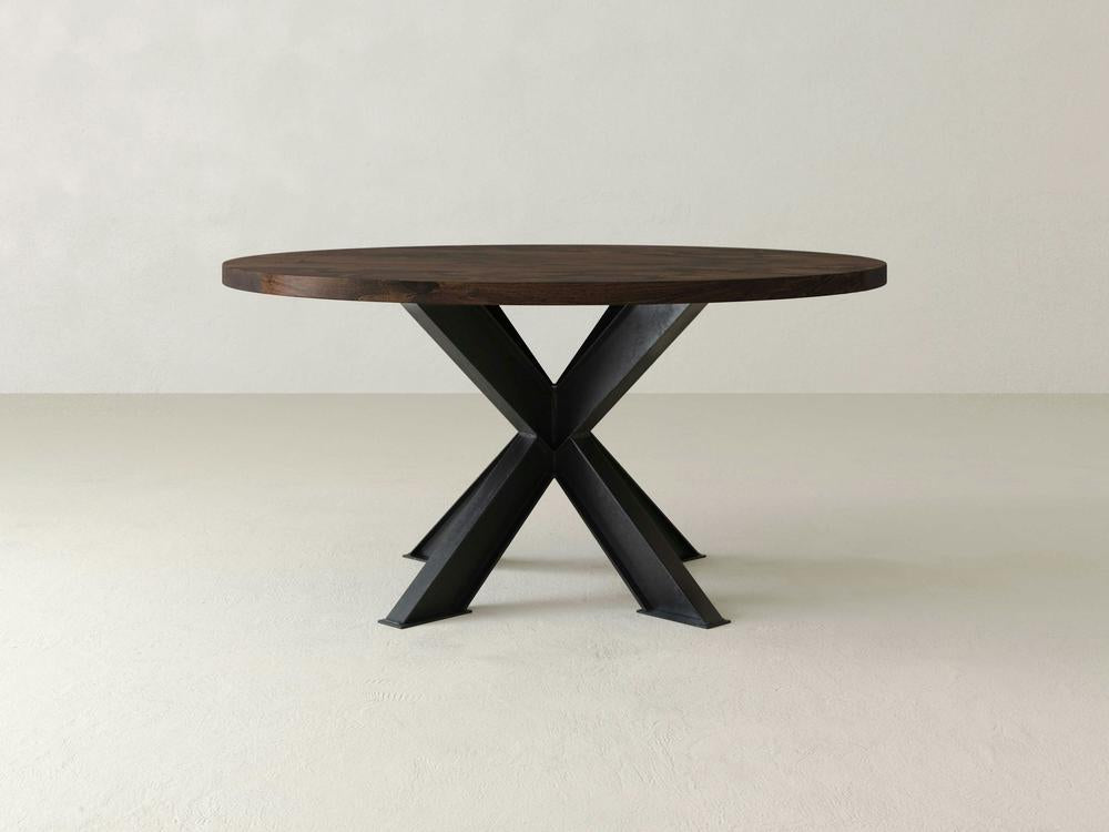 A Shiloh Round Dining Table - Tobacco by James+James, featuring a dark brown top and black X-shaped metal legs, is placed in a minimalistic room with a light-colored floor and wall.