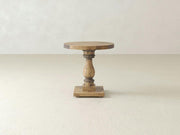 The Vivien Side Table - Harvest Wheat by James+James stands on a light-colored floor against a plain, light-colored wall. This rustic wooden pedestal table boasts a round top, a turned central support, and a square base. The natural and lightly weathered wood finish enhances its vintage charm.