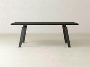 The Fulton Dining Table - Charred Ember by James+James boasts a minimalist, modern design featuring a rectangular dark wooden top and angled black metal legs, set against a plain, light-colored background. Its clean lines and sleek appearance ensure it fits seamlessly into any contemporary space.