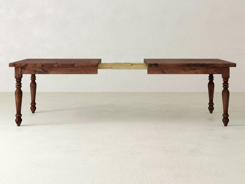 The James+James French Country Expandable Dining Table features a rich, dark wood top and ornate, turned legs. With its extension partially pulled out, the mechanism and additional leaves are revealed. The rustic beauty of this piece is highlighted against a plain, light-colored wall and floor.