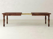 The James+James French Country Expandable Dining Table features a rich, dark wood top and ornate, turned legs. With its extension partially pulled out, the mechanism and additional leaves are revealed. The rustic beauty of this piece is highlighted against a plain, light-colored wall and floor.