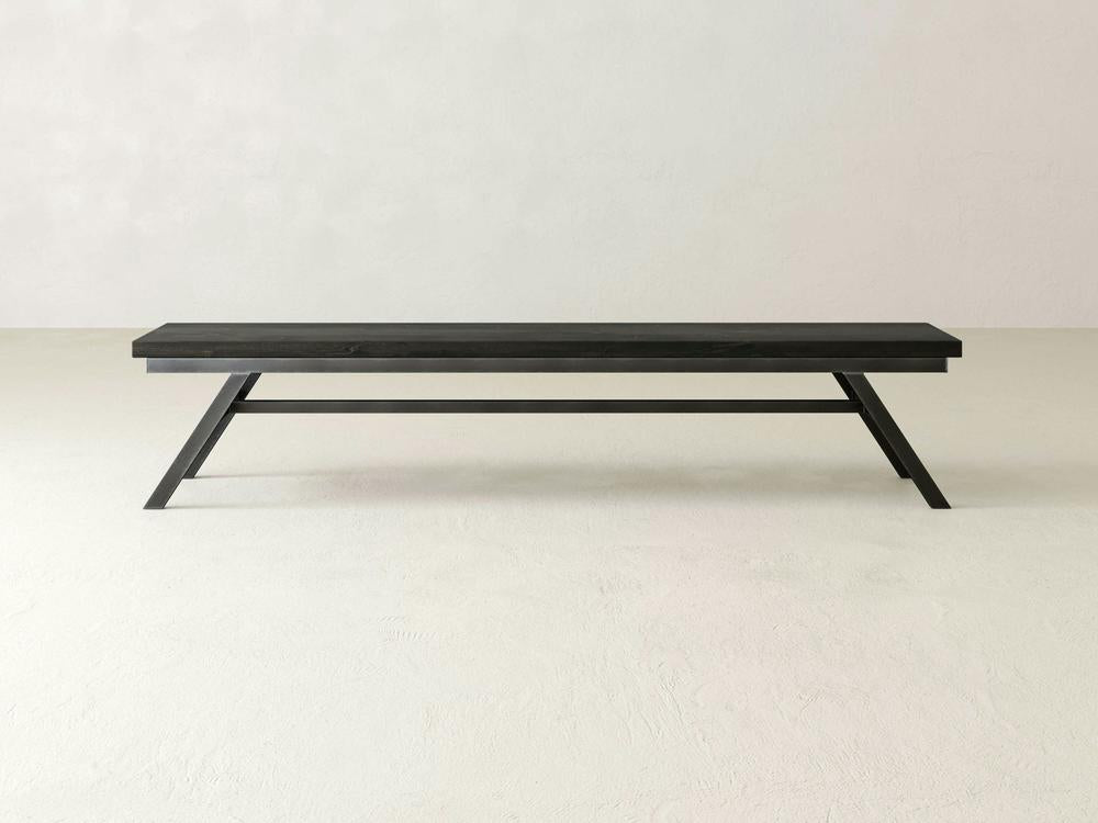 The James+James Fulton Bench in Charred Ember on Alder features a minimalist design with angled legs and a flat seat, shown against an off-white wall and light-colored floor.