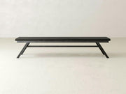 The James+James Fulton Bench in Charred Ember on Alder features a minimalist design with angled legs and a flat seat, shown against an off-white wall and light-colored floor.