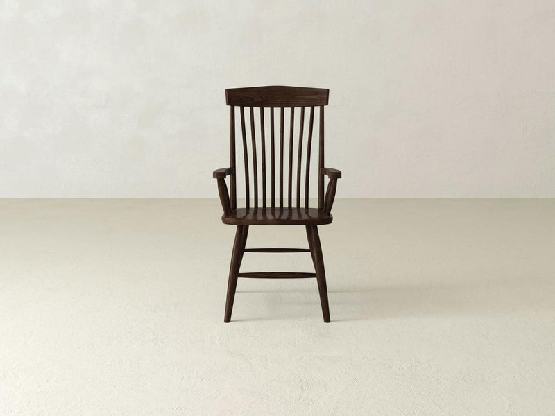 The Harper Arm Dining Chair by James & James showcases woodworking artistry in a minimalist room with light floors and walls. Crafted from dark wood, it features armrests and a high slatted backrest, offering a simple, classic design ideal for dining spaces.