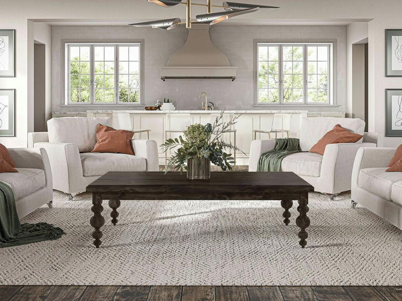 A cozy living room with a farmhouse style showcases two white, cushioned armchairs adorned with green and rust-colored throw pillows and blankets. The centerpiece is the Olivia Coffee Table in Charred Ember by James+James, standing proudly on a patterned rug. Two large windows frame the scene, with a glimpse of a white kitchen visible in the background.