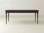 The Audrey Desk from James+James is a dark wooden table featuring four intricately carved legs, set against a plain, light-colored background. Its rectangular surface and two subtle front drawers enhance its timeless elegance, making it a classic addition to any space.