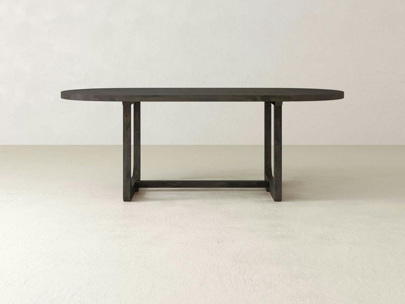 The James+James Emersen Oval Dining Table is crafted from knotty alder hardwood, showcasing a simplistic design with modern elegance. Its sturdy base features two vertical supports connected by a horizontal bar. The background highlights the table with light, neutral-colored walls and floors.
