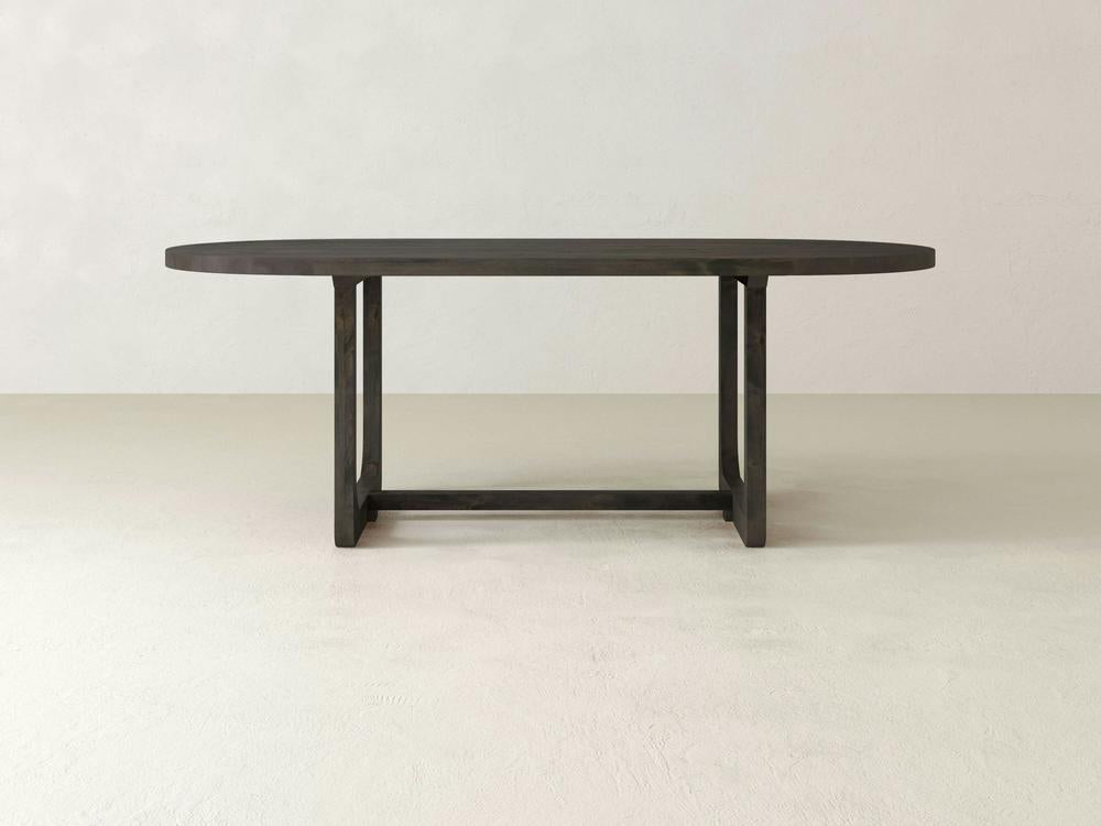 The James+James Emersen Oval Dining Table is crafted from knotty alder hardwood, showcasing a simplistic design with modern elegance. Its sturdy base features two vertical supports connected by a horizontal bar. The background highlights the table with light, neutral-colored walls and floors.