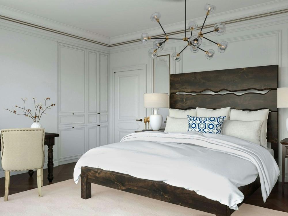 A modern bedroom featuring the James+James Live Edge Bed in Charred Ember on Alder, white bedding, a blue accent pillow, two bedside lamps, a desk with a cream chair, minimalist décor, and a contemporary chandelier.