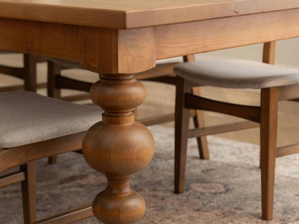 A close-up view of the Olivia Expandable Dining Table in Harvest Wheat by James+James, featuring intricately carved legs and matching wooden chairs with cushioned seats. The table and chairs are situated on a patterned rug, creating a warm and cozy ambiance.