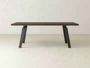 The Fulton Dining Table - Tobacco by James+James, featuring a dark brown rectangular wooden tabletop and black, angular metal legs, is placed in an empty room with light-colored walls and floor. The table boasts a sleek and modern design.