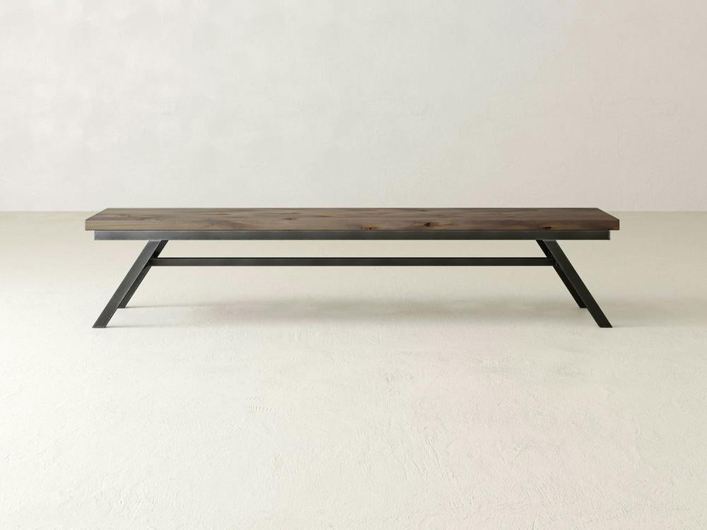 The Fulton Bench by James+James features a dark brown rectangular seat crafted from solid hardwood and black industrial steel bases that form angular supports. Placed against a plain light-colored wall on a smooth floor, it exemplifies a simple yet modern aesthetic.