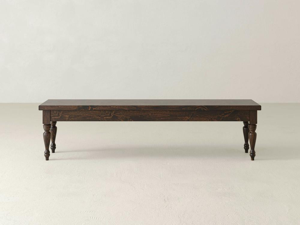 The James & James Chateau Bench - Tobacco features a simple, rustic design with dark wood and four turned legs, set against a plain, light-colored background.