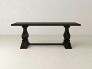 The Heirloom Dining Table - Charred Ember by James+James features a large, rectangular wooden top with a dark finish. It is supported by two thick, intricately carved pedestal legs connected by a horizontal stretcher. The table is set against a plain, white wall with a light-colored floor.