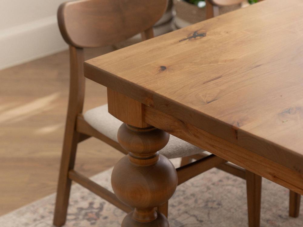 A close-up view of the Olivia Expandable Dining Table - Harvest Wheat from James+James, showcasing its polished surface and sturdy, carved legs. Behind the table is a matching wooden chair with a curved backrest and a cushioned seat. The furniture is placed on a patterned rug in a warmly lit room.