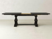 The Heirloom Expandable Dining Table - Charred Ember by James+James is a dark wooden dining table with two pedestal legs featuring intricate carving details. It has an extension mechanism in the center that, when partially extended, reveals a light-colored wood beneath. The background consists of a simple, light grey wall and floor.