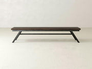The James+James Fulton Bench - Tobacco on Alder features a long, dark wood seat and angled black metal legs with a minimalist design, shown against a plain light-colored wall and floor.