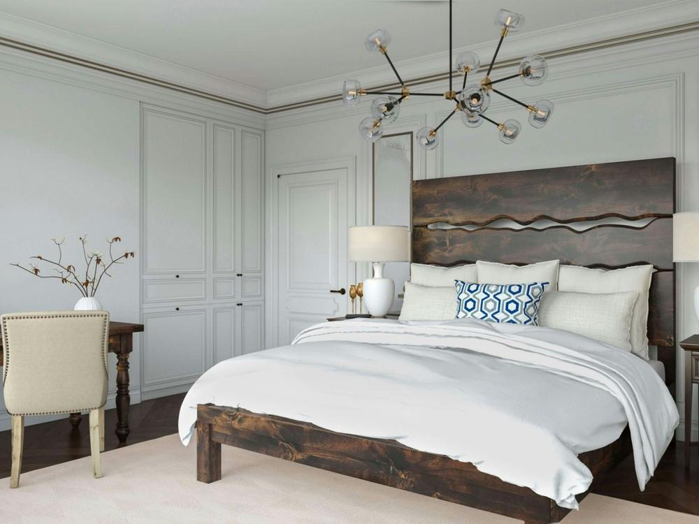 A modern bedroom features the James+James Live Edge Bed - Tobacco on Alder, white bedding with a blue accent pillow, two table lamps, a wooden desk with a beige chair, built-in white cabinets, and a contemporary chandelier.