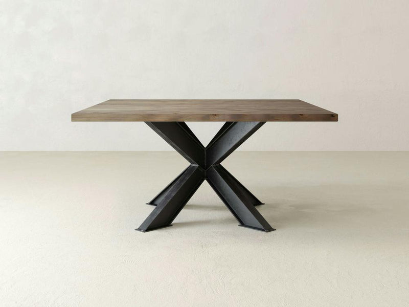 The James+James Shiloh Square Dining Table showcases a thick wooden top and a distinctive central metal base with four angled legs forming an X shape, perfectly capturing the essence of industrial chic against a plain, light-colored background.