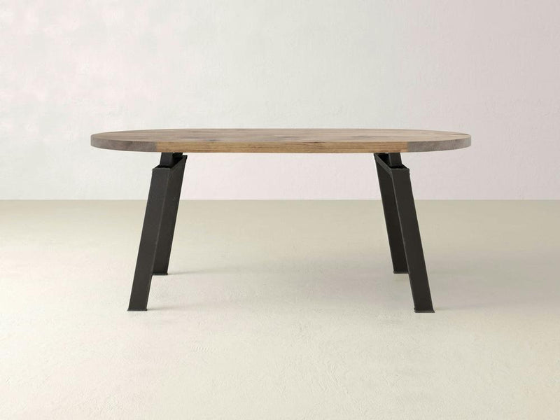 The Fulton Oval Dining Table by James+James features a modern industrial design with an oval, solid hardwood top and a smooth light brown surface. It is supported by four angled raw steel legs, creating a minimalist and contemporary look, all set against a neutral, light-colored background.