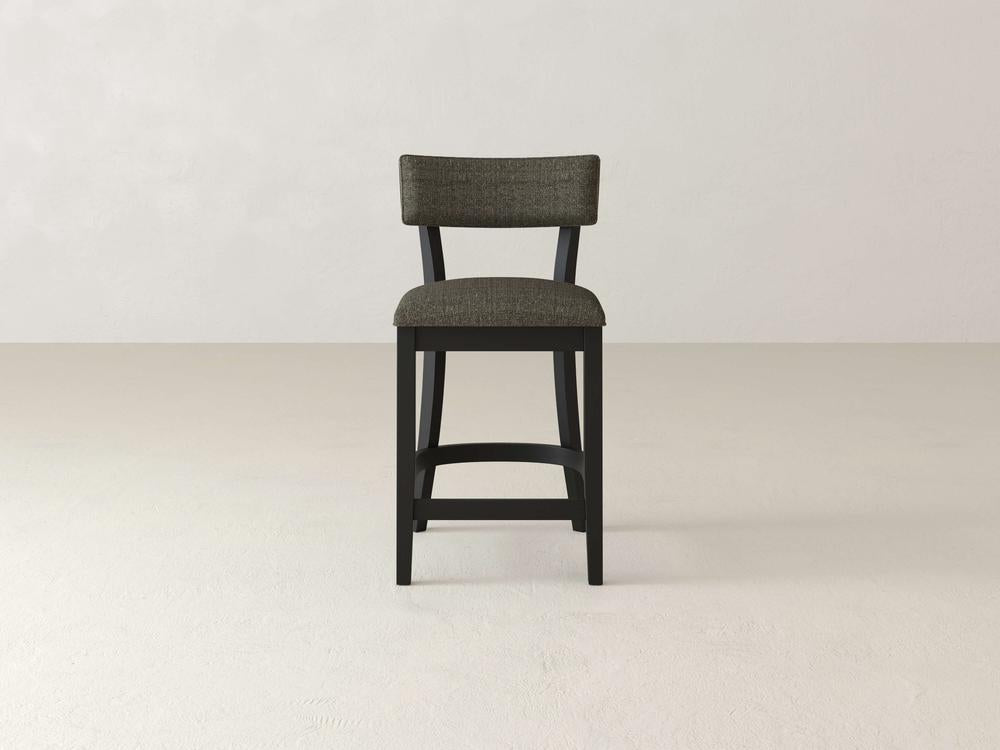 The Everly Upholstered Stool by James & James is a tall, dark wooden bar stool with a mid-century design, featuring a cushioned backrest and an upholstered seat in textured gray fabric, all highlighted against a light, neutral background.