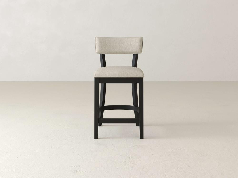 The Everly Upholstered Stool by James & James is a modern bar stool with a timeless style, featuring a beige upholstered seat and black wooden frame. Its design includes a rectangular backrest and straight legs with footrest, all against a light gray background with mid-century influences.