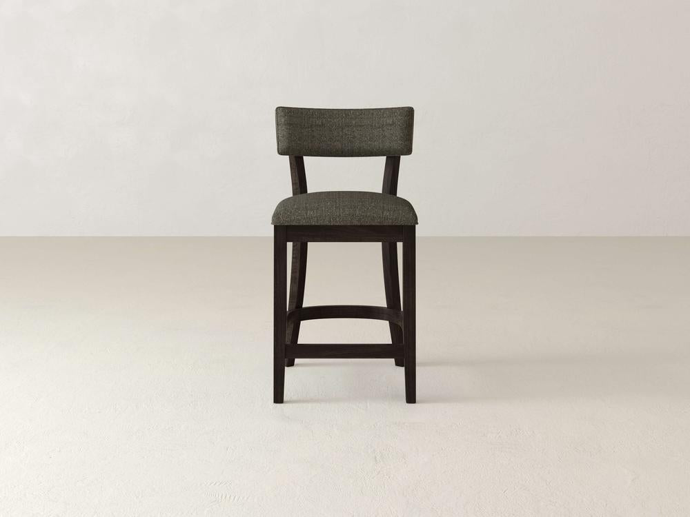The Everly Upholstered Stool by James & James features a wooden frame, padded backrest, and dark gray fabric seat, all set against a light gray background for a timeless look.