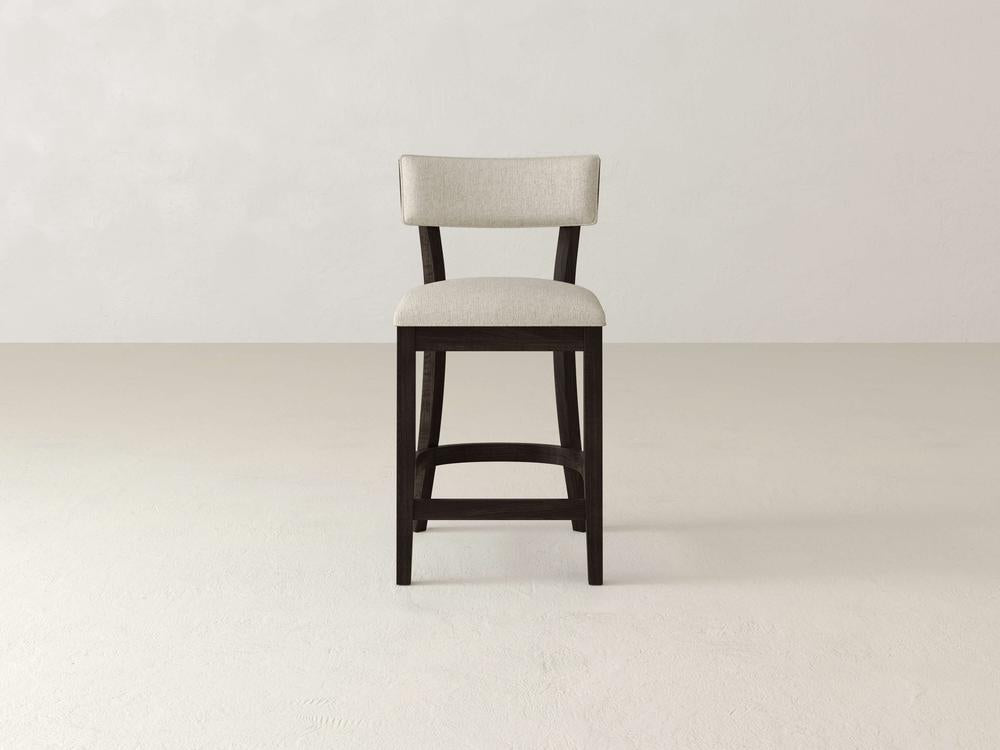The Everly Upholstered Stool by James & James, featuring a beige seat and backrest, stands on a light floor against an off-white wall. This mid-century design piece with a dark frame is perfectly centered in the minimalist space.