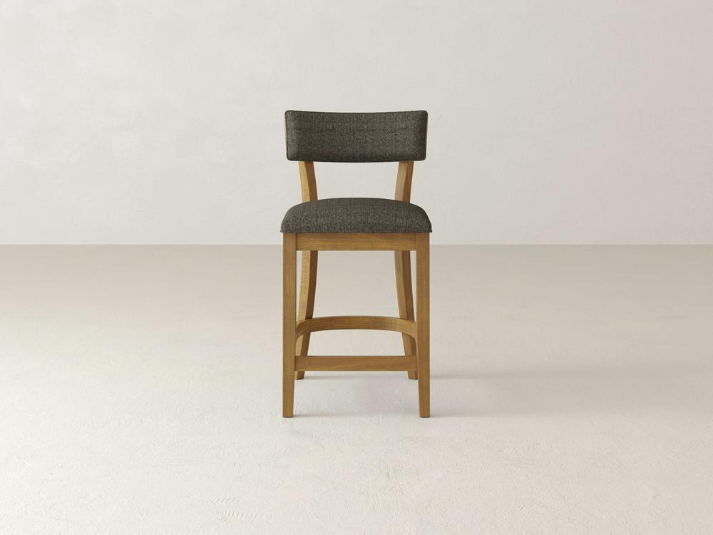 The Everly Upholstered Stool by James & James, featuring a mid-century wooden design and a cushioned seat in dark gray fabric, stands elegantly against a plain, light-colored background.