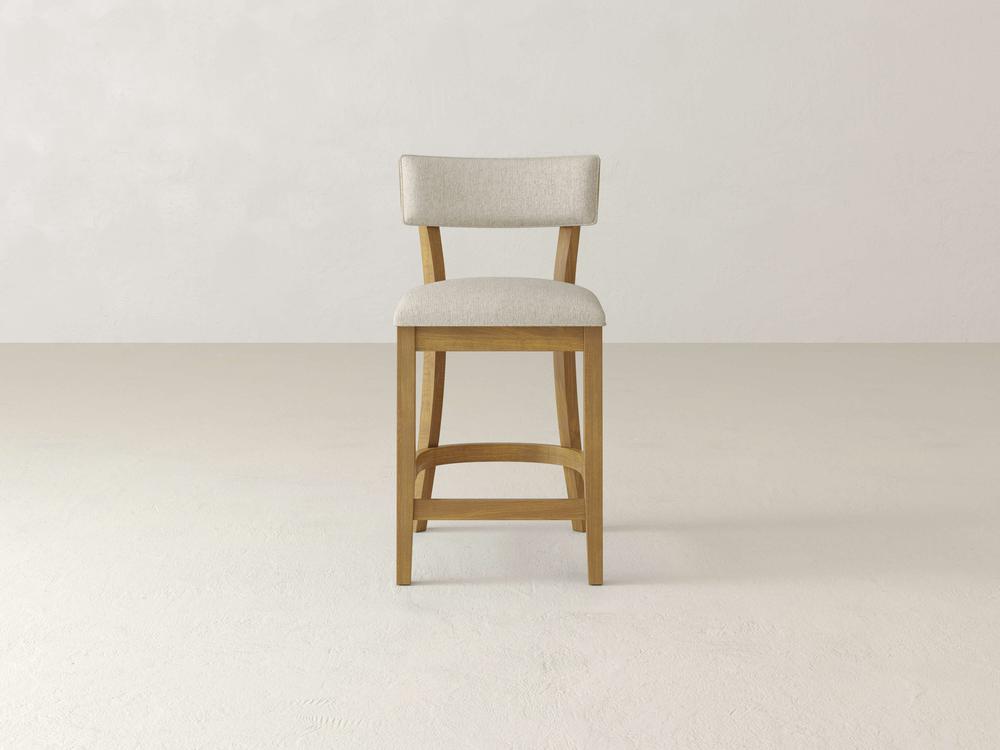 The Everly Upholstered Stool by James & James, featuring a wooden frame with a light beige seat and backrest, brings timeless style to any minimalist space set against neutral tones.