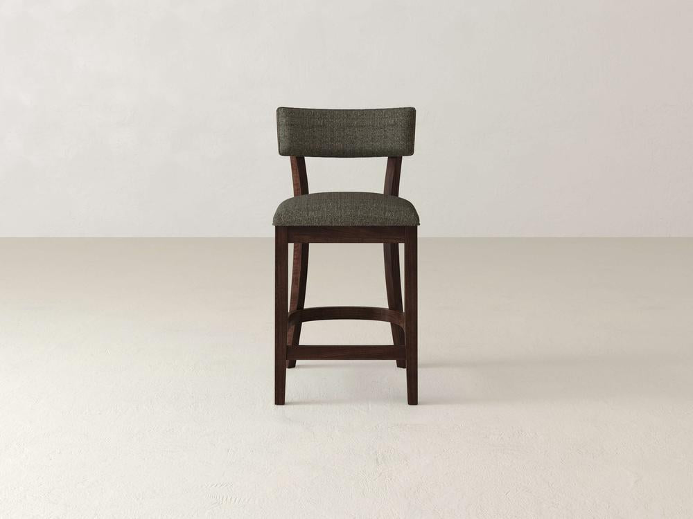 The Everly Upholstered Stool by James & James features a wooden frame, padded backrest, and upholstered seat in dark gray fabric. Set against a light background, it blends classic and modern design for a timeless mid-century appeal.