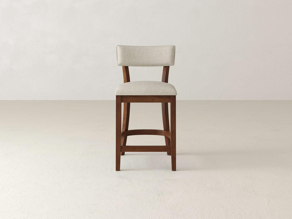 The Everly Upholstered Stool by James & James is a tall wooden bar stool with a light beige cushioned seat and backrest, embodying timeless style against a plain, light-colored background.