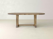 The Thaden Oval Dining Table - Harvest Wheat by James+James features a light wooden design with sturdy legs and a crossbar. It is set against a plain, light-colored wall and floor background, showcasing its simple, rustic aesthetic with a smooth surface and visible wood grain.