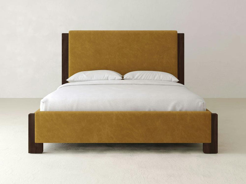 The James & James Lennon Bed – Velvet features a luxurious mustard yellow velvet upholstered headboard and footboard, dark wood frame with maple accents, complemented by white bedding and two pillows, all set in a minimalist room with light-colored walls and floor.