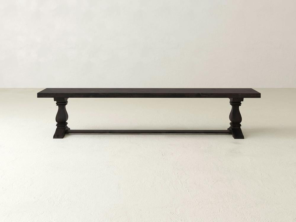 The James+James Heirloom Bench in Charred Ember on Alder features a rectangular seat and carved legs, set on a light floor against an off-white wall.