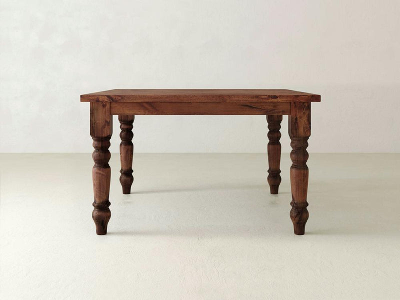 The Baluster Square Dining Table - Tuscany by James+James features a rectangular top and four intricately carved legs, set in a minimalist environment with a light-colored floor and a white wall backdrop. The table boasts a rustic look with its natural wood finish.