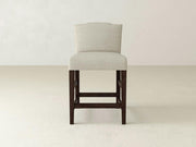 The Hadley Upholstered Stool by James & James features beige fabric, cushioned seat and backrest, sleek dark wooden legs, and a contemporary design that enhances any dining space against a light background.