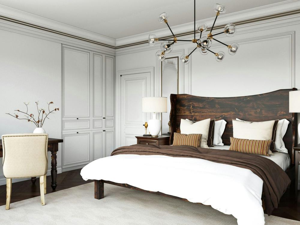 A stylish bedroom featuring the Harlow Bed in Tobacco on Alder by James+James, white and brown bedding, a modern chandelier, white walls with molding, built-in cabinets, a desk with a beige chair, and decorative branches in a vase.